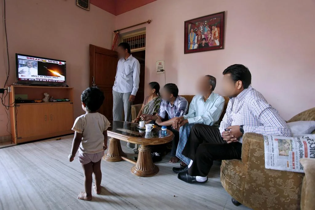 A family in a living room in India watching a news channel on a single TV, with a toddler standing in front of the screen